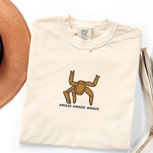 May include: A cream-colored short-sleeve t-shirt featuring a brown, multi-legged creature graphic with green accents. The creature is making peace signs with two of its limbs. Below the graphic, the text reads AMAZE AMAZE AMAZE in black letters.