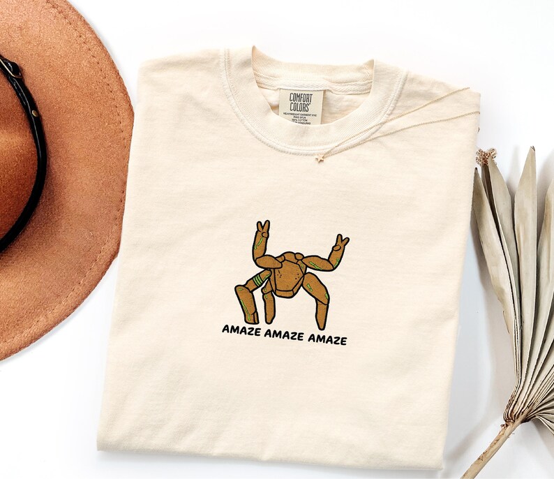 May include: A cream-colored short-sleeve t-shirt featuring a brown, multi-legged creature graphic with green accents. The creature is making peace signs with two of its limbs. Below the graphic, the text reads AMAZE AMAZE AMAZE in black letters.