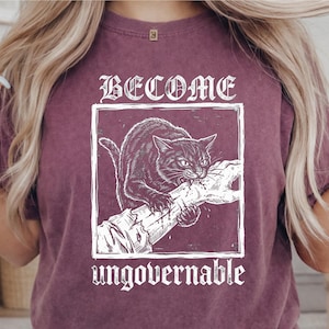 May include: A maroon short-sleeve t-shirt featuring a white graphic print of a cat biting a hand inside a square frame. The text BECOME is printed above the frame and the word ungovernable is printed below the frame in a white gothic font.