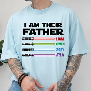 May include: Light blue t-shirt with the text "I AM THEIR FATHER" in black. Below the text are four lightsabers with the names Liam, Owen, Zoey, and Ayla in red, green, blue, and purple.