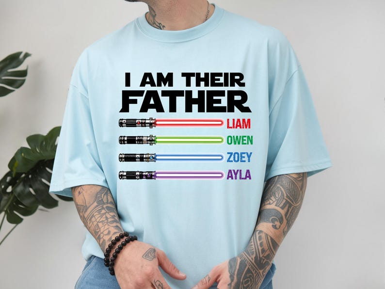 May include: Light blue t-shirt with the text "I AM THEIR FATHER" in black. Below the text are four lightsabers with the names Liam, Owen, Zoey, and Ayla in red, green, blue, and purple.