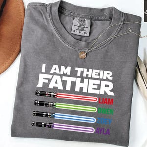 May include: A gray Comfort Colors t-shirt with the text "I AM THEIR FATHER" in white. Below the text are four lightsabers in red, green, blue, and purple, with names Liam, Owen, Joey, and Ayla. A silver necklace is draped on the shirt.