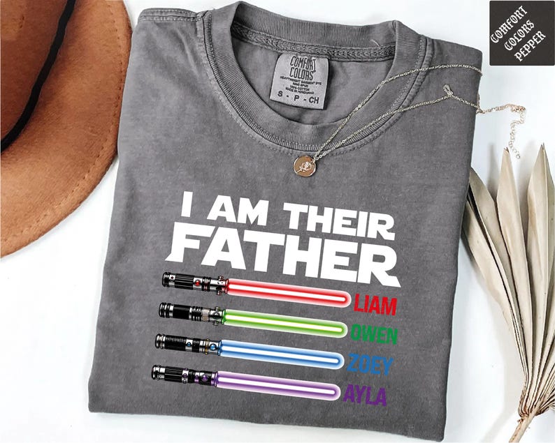 May include: A gray Comfort Colors t-shirt with the text "I AM THEIR FATHER" in white. Below the text are four lightsabers in red, green, blue, and purple, with names Liam, Owen, Joey, and Ayla. A silver necklace is draped on the shirt.