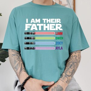 May include: Teal t-shirt with the text "I AM THEIR FATHER" in white. Below the text are four lightsabers in red, green, blue, and purple, each with a name: Liam, Owen, Zoey, and Ayla. The shirt is a casual, short-sleeved design.