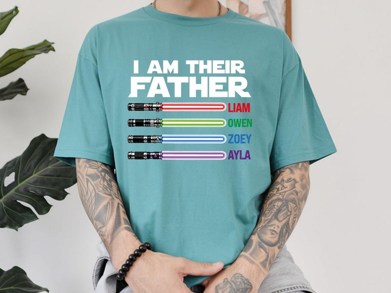May include: Teal t-shirt with the text "I AM THEIR FATHER" in white. Below the text are four lightsabers in red, green, blue, and purple, each with a name: Liam, Owen, Zoey, and Ayla. The shirt is a casual, short-sleeved design.
