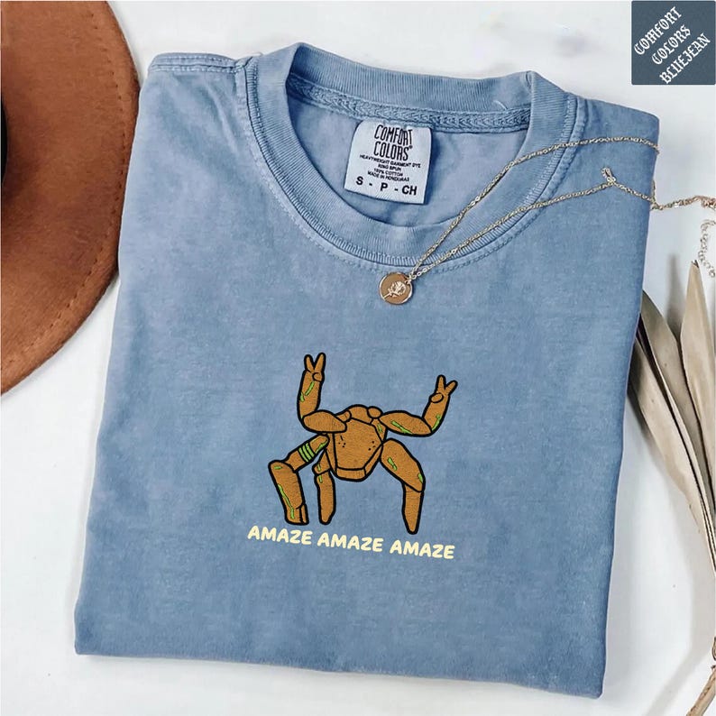 May include: A blue short-sleeve t-shirt featuring a graphic of a brown, multi-legged creature with green accents and the text "AMAZE AMAZE AMAZE" printed below. The shirt has a crew neckline and a Comfort Colors brand label on the inside collar.