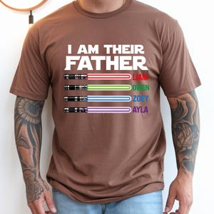 May include: A brown t-shirt with the text "I AM THEIR FATHER" and four colored lightsabers. Each lightsaber has a name: Liam, Owen, Zoey, and Ayla. The lightsabers are red, green, blue, and purple.
