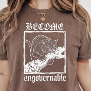 May include: A brown short-sleeved t-shirt featuring a white graphic print of a cat biting a hand. The text BECOME is printed above the cat, and the word ungovernable is printed below the graphic in a stylized, distressed white font.