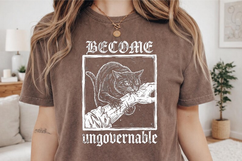 May include: A brown short-sleeved t-shirt featuring a white graphic print of a cat biting a hand. The text BECOME is printed above the cat, and the word ungovernable is printed below the graphic in a stylized, distressed white font.