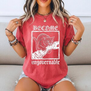 May include: A red short-sleeve t-shirt featuring a white graphic of a cat biting a hand inside a square frame. The text BECOME is printed above the frame and the word ungovernable is printed below the frame in a white distressed font.