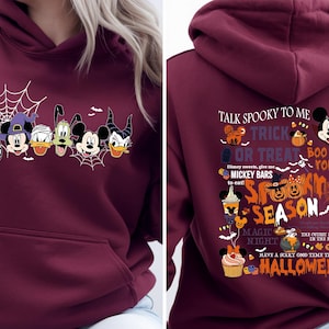 May include: A maroon hooded sweatshirt featuring a front graphic of cartoon characters in Halloween costumes and a back graphic with the text Talk Spooky To Me, Trick Or Treat, Boo To You, Mickey Bars, Spooky Season, Magic Night, and Halloween.