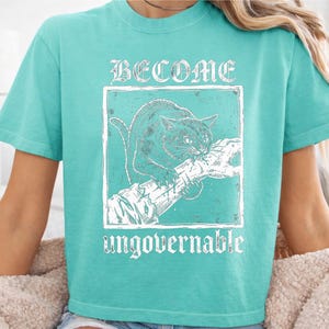 May include: A bright turquoise short-sleeve t-shirt featuring a white graphic print of a cat biting a branch. The text BECOME is printed above the cat, and the word ungovernable is printed below the cat in a distressed, weathered font style.