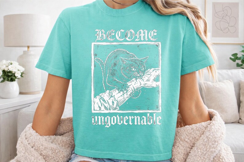 May include: A bright turquoise short-sleeve t-shirt featuring a white graphic print of a cat biting a branch. The text BECOME is printed above the cat, and the word ungovernable is printed below the cat in a distressed, weathered font style.