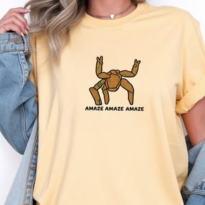 May include: A pale yellow short-sleeved t-shirt featuring a graphic of a brown, multi-legged creature with green accents making peace signs with its upper limbs. Below the creature, the text AMAZE AMAZE AMAZE is printed in black capital letters.
