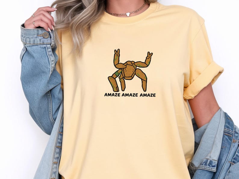 May include: A pale yellow short-sleeved t-shirt featuring a graphic of a brown, multi-legged creature with green accents making peace signs with its upper limbs. Below the creature, the text AMAZE AMAZE AMAZE is printed in black capital letters.