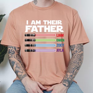 May include: A peach-colored t-shirt with the text "I AM THEIR FATHER" in white. Below the text are four lightsabers with the names Liam, Owen, Zoey, and Ayla. The lightsabers have red, green, blue, and purple blades.