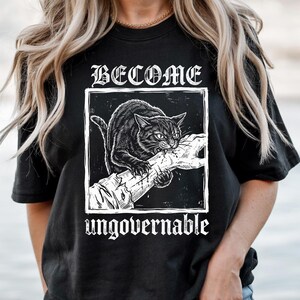 May include: A black t-shirt featuring a white graphic print of a cat biting a human arm. The text BECOME is printed in a gothic font above the square illustration, and the word ungovernable is printed in a matching gothic font below the image.