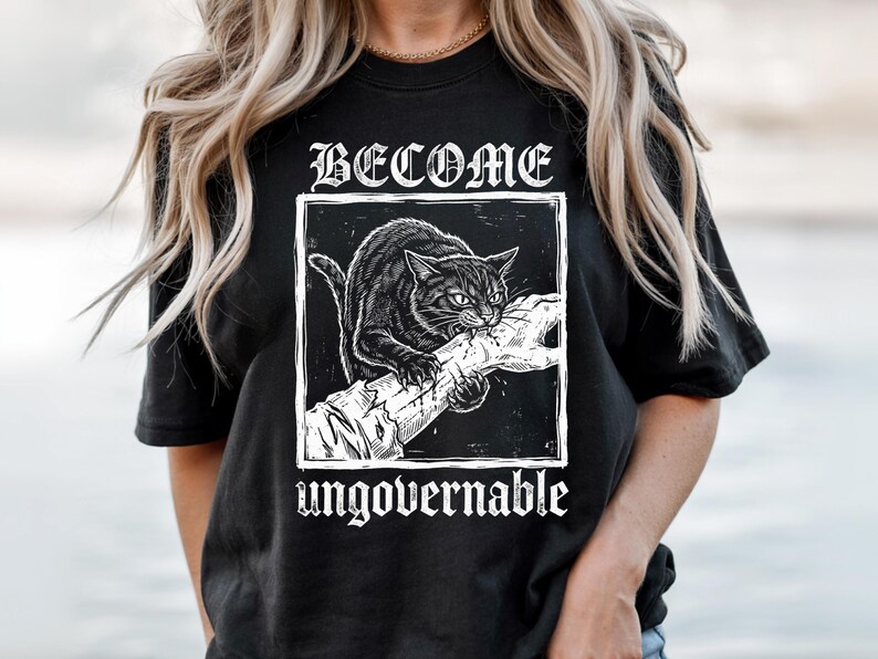 May include: A black t-shirt featuring a white graphic print of a cat biting a human arm. The text BECOME is printed in a gothic font above the square illustration, and the word ungovernable is printed in a matching gothic font below the image.