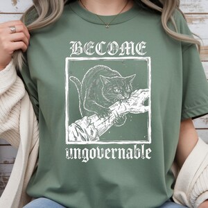 May include: A sage green t-shirt featuring a white graphic print of a cat biting a human hand inside a square frame. The text BECOME is printed above the frame and the word ungovernable is printed below the frame in a distressed white font.