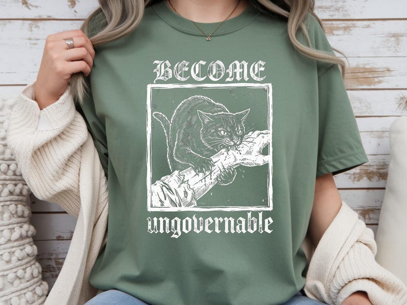 May include: A sage green t-shirt featuring a white graphic print of a cat biting a human hand inside a square frame. The text BECOME is printed above the frame and the word ungovernable is printed below the frame in a distressed white font.