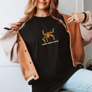May include: A black crewneck t-shirt featuring a gold-colored graphic of a multi-legged creature with the text "AMAZE AMAZE AMAZE" printed in gold underneath. The shirt is worn under a tan and cream-colored button-up varsity-style jacket.