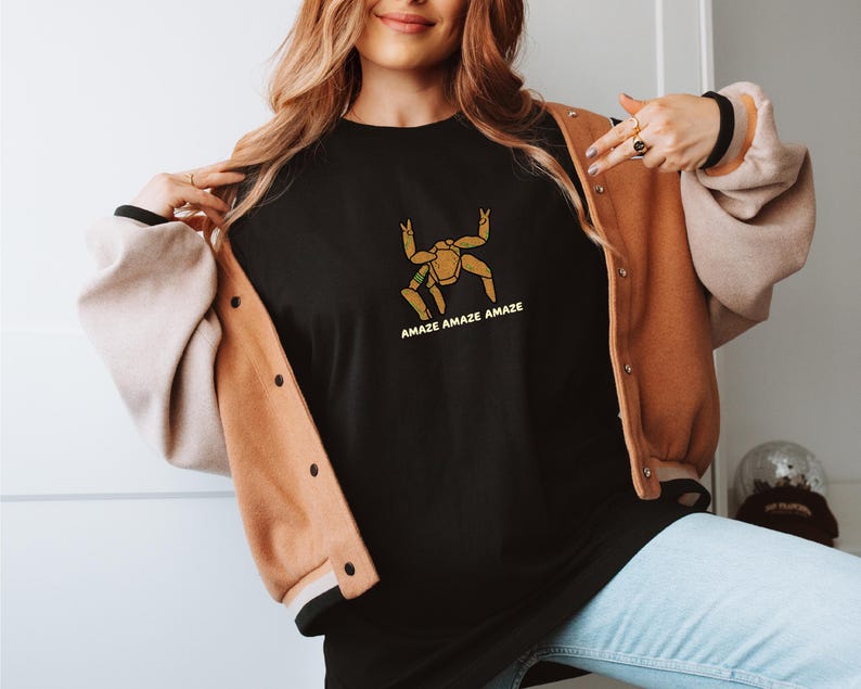 May include: A black crewneck t-shirt featuring a gold-colored graphic of a multi-legged creature with the text "AMAZE AMAZE AMAZE" printed in gold underneath. The shirt is worn under a tan and cream-colored button-up varsity-style jacket.