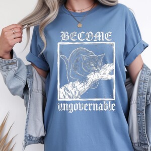 May include: A blue t-shirt featuring a white graphic print of a cat biting a tree branch inside a square frame. The text "BECOME" is printed in a gothic font above the frame, and the word "ungovernable" is printed in a matching font below it.