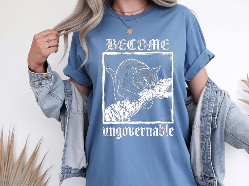 May include: A blue t-shirt featuring a white graphic print of a cat biting a tree branch inside a square frame. The text "BECOME" is printed in a gothic font above the frame, and the word "ungovernable" is printed in a matching font below it.