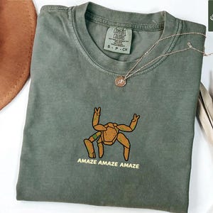 May include: A moss green short-sleeved t-shirt featuring an embroidered graphic of a tan, multi-legged creature with green accents. Below the creature, the text reads "AMAZE AMAZE AMAZE" in yellow. The shirt has a Comfort Colors brand label at the neck.