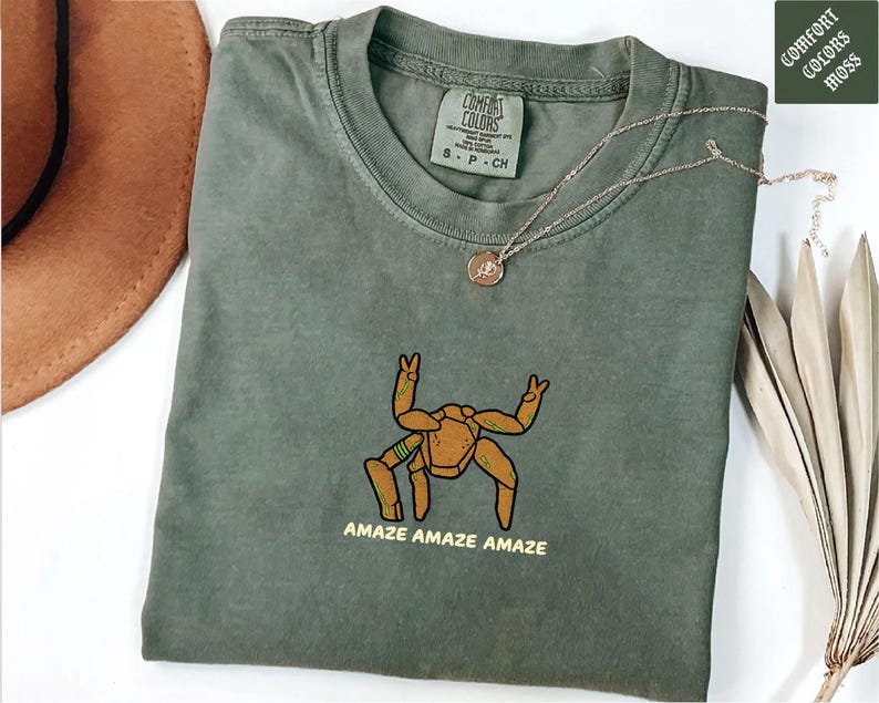 May include: A moss green short-sleeved t-shirt featuring an embroidered graphic of a tan, multi-legged creature with green accents. Below the creature, the text reads "AMAZE AMAZE AMAZE" in yellow. The shirt has a Comfort Colors brand label at the neck.