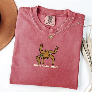 May include: A dusty rose t-shirt featuring a graphic of a brown, multi-legged creature with raised arms and the text "AMAZE AMAZE AMAZE" printed in yellow. The shirt has a Comfort Colors brand label at the neck and a crew neckline design.