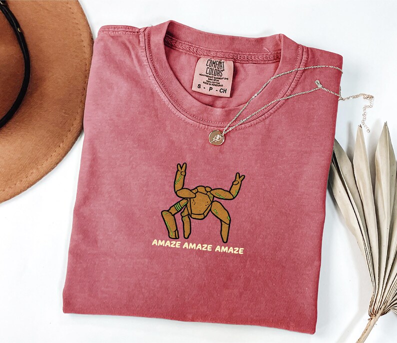 May include: A dusty rose t-shirt featuring a graphic of a brown, multi-legged creature with raised arms and the text "AMAZE AMAZE AMAZE" printed in yellow. The shirt has a Comfort Colors brand label at the neck and a crew neckline design.