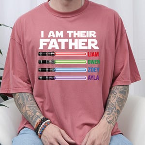 May include: A dusty rose-colored t-shirt with the text "I AM THEIR FATHER" in white block letters. Below the text are four lightsabers with the names Liam, Owen, Zoey, and Ayla in red, green, blue, and purple.