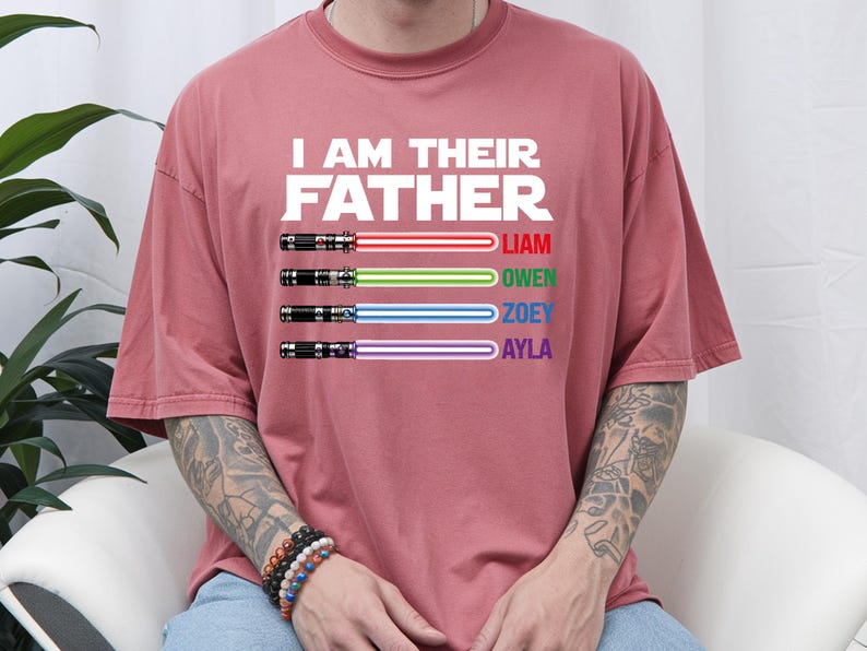 May include: A dusty rose-colored t-shirt with the text "I AM THEIR FATHER" in white block letters. Below the text are four lightsabers with the names Liam, Owen, Zoey, and Ayla in red, green, blue, and purple.