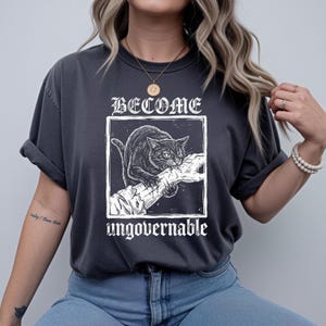 May include: A charcoal gray t-shirt featuring a white graphic print of a cat biting a hand inside a square frame. The text BECOME is printed above the frame and the word ungovernable is printed below the frame in a stylized white font.