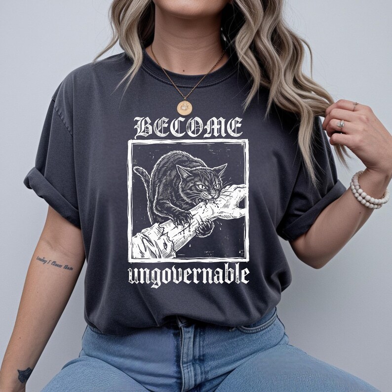 May include: A charcoal gray t-shirt featuring a white graphic print of a cat biting a hand inside a square frame. The text BECOME is printed above the frame and the word ungovernable is printed below the frame in a stylized white font.
