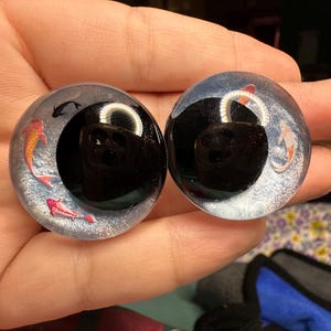 May include: Two round, clear glass eyes with black pupils and a design of orange and pink fish swimming inside. The eyes are on a hand.