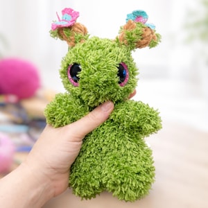 May include: A green plush toy made of fuzzy yarn with large, dark, glittery eyes. The creature features two tan, textured protrusions on its head, each topped with a small, colorful crocheted butterfly in shades of pink and light blue.