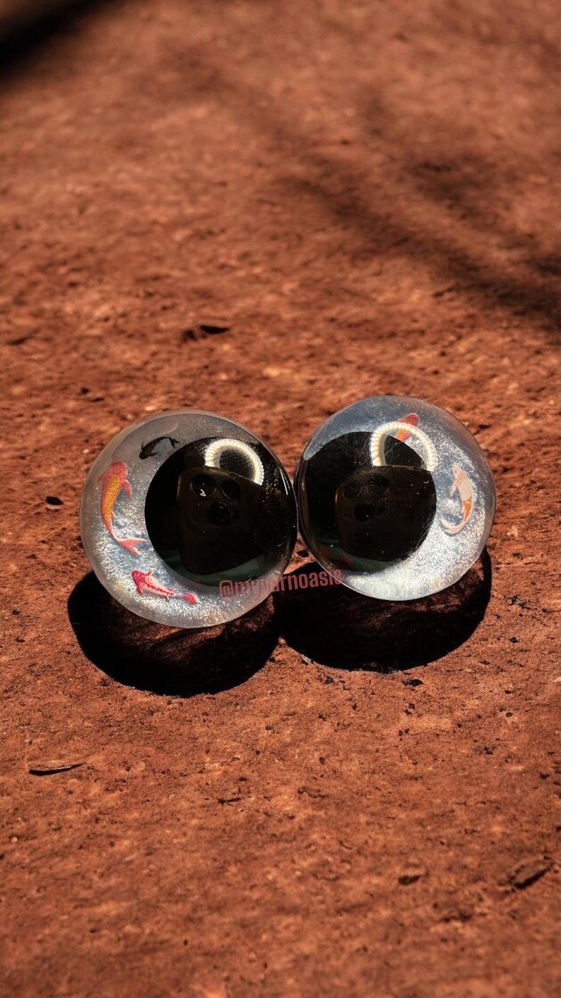 May include: A pair of circular ear plugs made of clear resin with embedded glitter and small koi fish illustrations. Each plug features a large black circular inlay in the centre and the text @myoarnoasis printed in small white letters on the bottom.