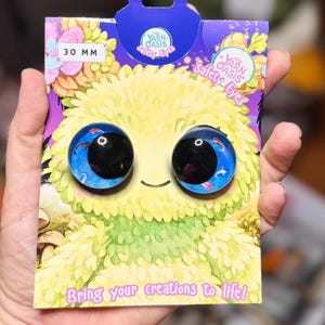 May include: A pair of 30 mm round safety eyes for crafts, featuring a blue background with small fish illustrations and large black pupils. The eyes are mounted on a purple and yellow illustrated display card labelled My Yarn Oasis Safety Eyes.