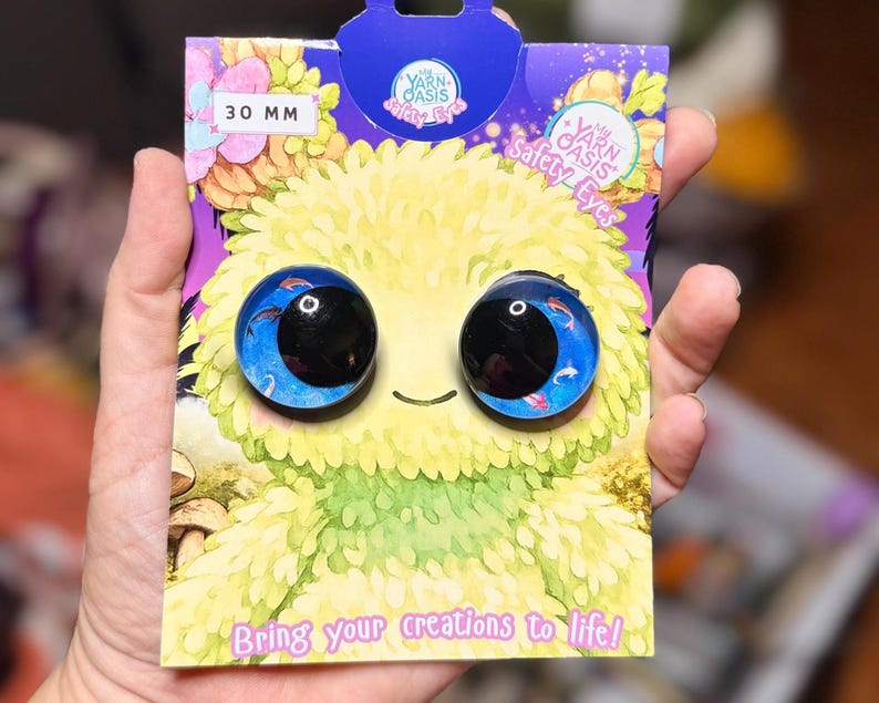 May include: A pair of 30 mm round safety eyes for crafts, featuring a blue background with small fish illustrations and large black pupils. The eyes are mounted on a purple and yellow illustrated display card labelled My Yarn Oasis Safety Eyes.