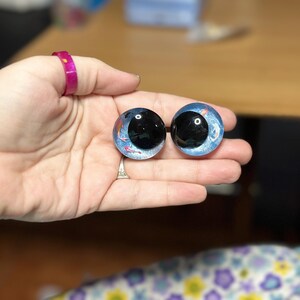 May include: Two round, glass-like cabochons featuring a blue, glittery background with small, painted orange and white koi fish motifs. Each piece has a large, glossy black circular centre, resembling a realistic eye with a deep, reflective finish.