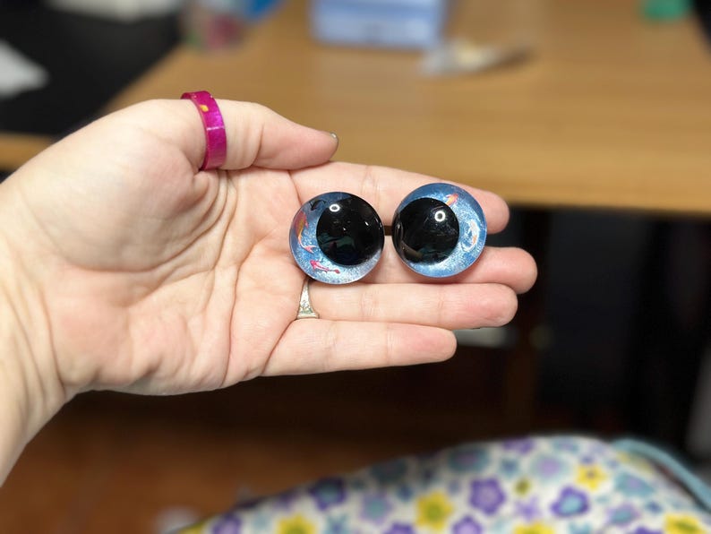 May include: Two round, glass-like cabochons featuring a blue, glittery background with small, painted orange and white koi fish motifs. Each piece has a large, glossy black circular centre, resembling a realistic eye with a deep, reflective finish.