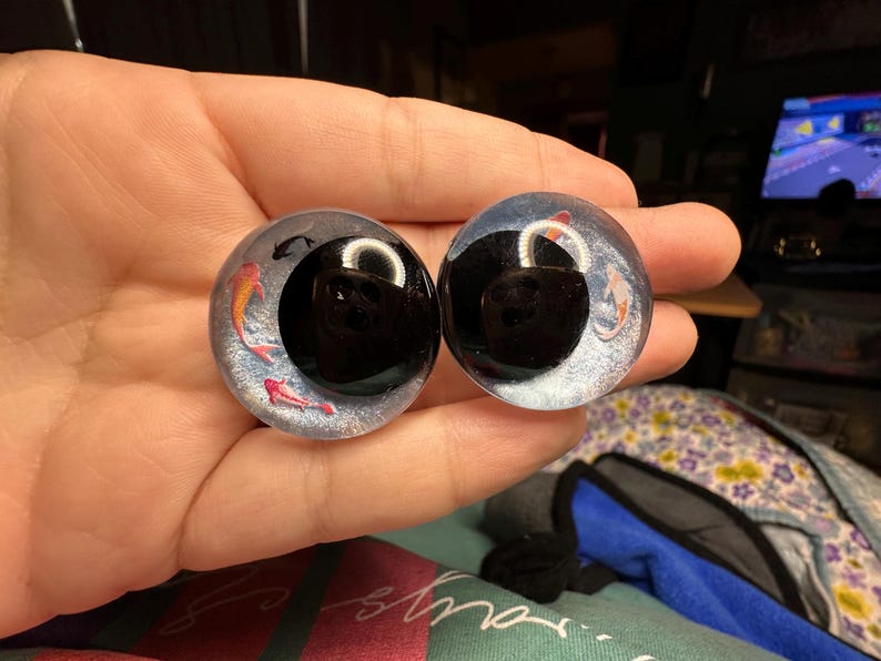 May include: Two round, clear glass eyes with black pupils and a design of orange and pink fish swimming inside. The eyes are on a hand.