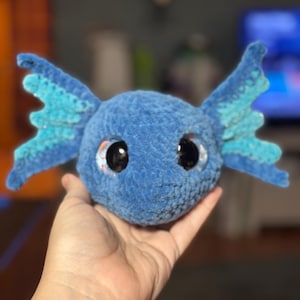 May include: A blue crocheted plush toy shaped like an axolotl head with large black eyes and prominent, textured side fins. The plush features a soft, fuzzy yarn texture in a deep blue shade, with lighter blue accents on the wide, fan-like fins.