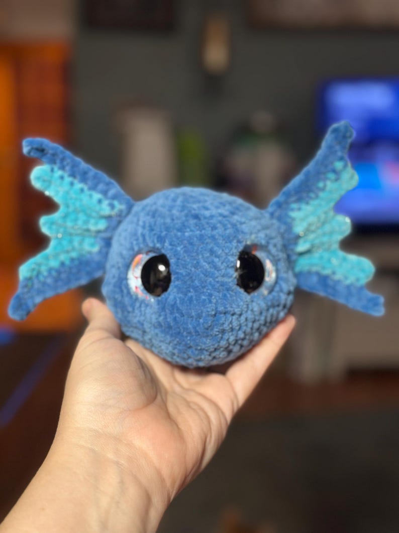 May include: A blue crocheted plush toy shaped like an axolotl head with large black eyes and prominent, textured side fins. The plush features a soft, fuzzy yarn texture in a deep blue shade, with lighter blue accents on the wide, fan-like fins.