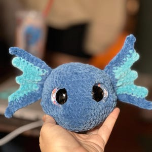 May include: A round, blue, crocheted plush toy featuring large, glossy black eyes and two prominent, wing-like side fins in shades of light and dark blue. The texture of the yarn is soft and velvety, creating a rounded head shape with detailed, textured fins extending from each side.