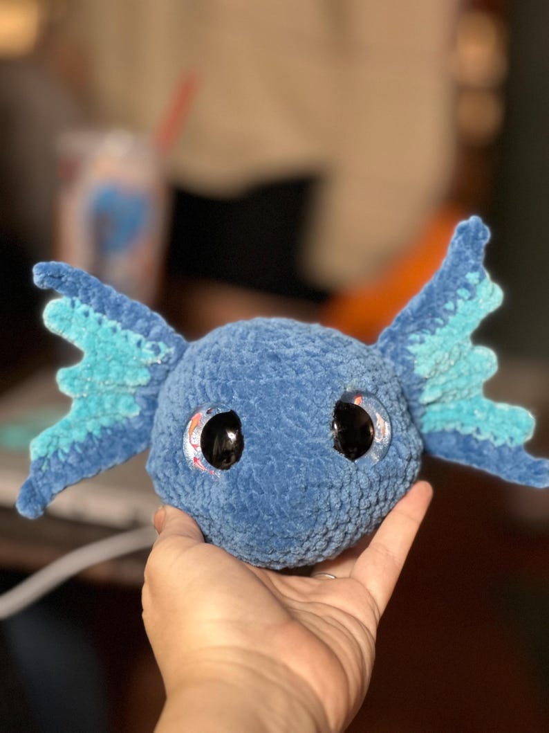 May include: A round, blue, crocheted plush toy featuring large, glossy black eyes and two prominent, wing-like side fins in shades of light and dark blue. The texture of the yarn is soft and velvety, creating a rounded head shape with detailed, textured fins extending from each side.