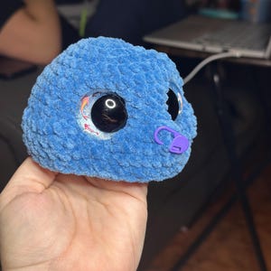 May include: A round, blue, textured crochet plush toy featuring large, glossy black safety eyes with a subtle koi fish graphic inside the iris. A small, purple plastic stitch marker is attached to the side of the blue yarn surface.