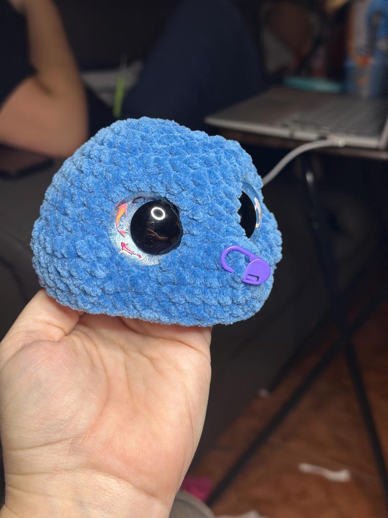 May include: A round, blue, textured crochet plush toy featuring large, glossy black safety eyes with a subtle koi fish graphic inside the iris. A small, purple plastic stitch marker is attached to the side of the blue yarn surface.